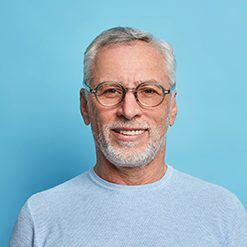 Portrait of handsome bearded European man with grey hair and beard smiles pleasantly looks directly at camera being in good mood has lucky day wears spectacles and sweater isolated over blue wall
